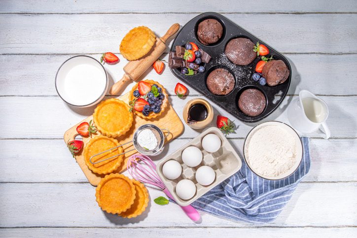 Baking Ingredients and supplies surrounding a muffin pan with chocolate muffins in it an small tarts with berries on them